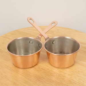 May include: Two small copper sauce pans with handles. The pans are sitting on a wooden surface.