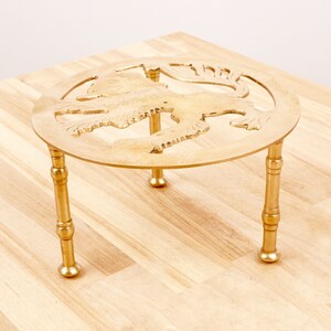 Puede incluir: Un salvamanteles de metal dorado con una parte superior redonda y tres patas. La parte superior presenta un diseño decorativo de un pájaro estilizado.