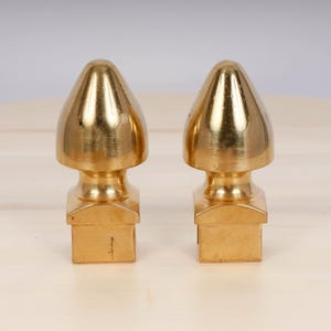 Op de afbeelding: Twee gouden metalen eindstukken met een vierkante basis en een puntige top.