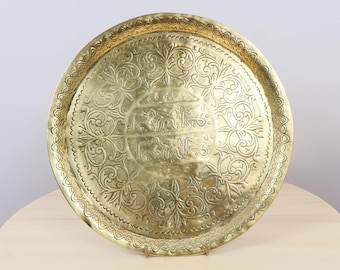 May include: A round, antique brass tray with intricate floral and script engravings. The tray has a scalloped edge and a polished finish, resting on a small stand. The design features detailed patterns and a central inscription, showcasing a vintage aesthetic.