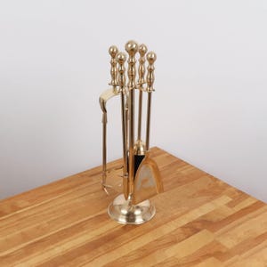 May include: A brass fireplace tool set with a stand. The set includes a poker, shovel, brush, and tongs. The tools have decorative ball finials and are arranged on a wooden surface.