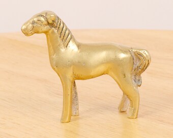May include: A small, gold-colored brass horse figurine. The horse is standing on all four legs with its head held high. The figurine is a simple design with no intricate details.