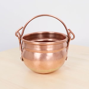 Puede incluir: Un pequeño caldero de color cobre con un cuerpo redondeado y un asa curva. El caldero tiene un acabado brillante y está diseñado con un asa para facilitar su transporte. El diseño general es simple y clásico.