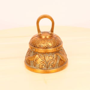 Puede incluir: Una pequeña campana de latón adornada con un asa. La campana está decorada con intrincados tallados de hojas y animales.