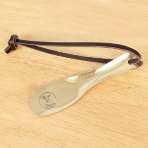 May include: A brass shoe horn with a brown leather strap. The shoe horn has a rounded end and is engraved with the words "Wear X Shoes".