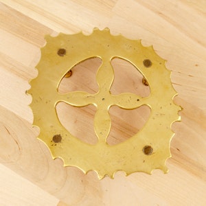 May include: A brass trivet with a decorative, four-leaf clover design. The trivet has a scalloped edge and four small feet.