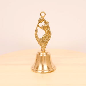 May include: A vintage brass bell with a detailed cherub figure on the handle. The bell has a classic shape with a flared base and a loop for hanging. The cherub is holding a floral element. The bell is displayed on a light wooden surface.