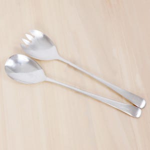 May include: A pair of silver-toned metal salad serving utensils consisting of one solid rounded spoon and one matching spoon with three distinct tines at the top edge. Both utensils feature long, smooth, slender handles and a polished finish.