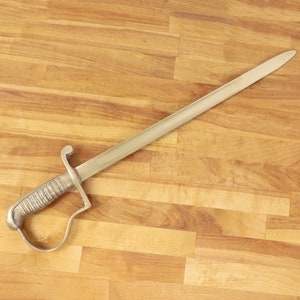 May include: A gold-colored sword with a decorative handle and a long, straight blade. The sword is lying on a wooden surface.