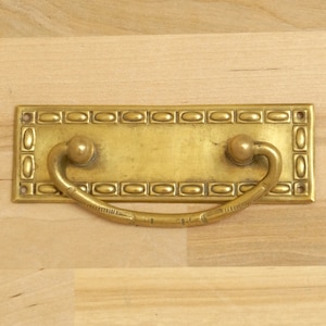 May include: A rectangular brass drawer pull with an oval pattern border and a curved handle. The handle has a ball on each end. The brass has a slightly aged appearance, suggesting vintage or antique origins. The hardware is designed for furniture.