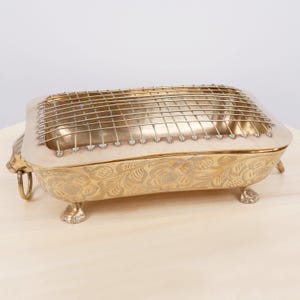 Peut inclure: Un plat de service rectangulaire en laiton avec une gravure florale complexe sur les côtés, quatre pieds en forme de griffe et une grille métallique amovible sur le dessus. Le plat comprend une petite poignée annulaire et un lion décoratif.
