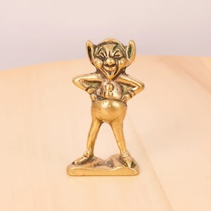 以下が含まれることがあります： 尖った耳といたずらっぽい表情をした笑顔のノームの金色の金属製フィギュア。ノームは手を腰に当てて立っており、シンプルな服装をしています。フィギュアは小さな平らな台座の上に置かれています。