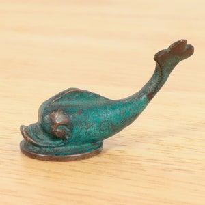 May include: A small, teal-colored metal dolphin figurine with a rounded base. The dolphin is curled into a playful pose with its tail raised.
