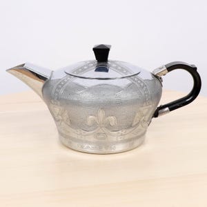 Puede incluir: Una tetera plateada con asa y tapa negras. La tetera tiene un diseño ornamentado y la palabra "ELIZABETH" está grabada en la base. El pico está en ángulo y el asa es curva.