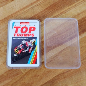 Puede incluir: Un mazo de cartas "Super Top Trumps Racing Bikes" en una funda de plástico transparente. El diseño de la carta presenta un corredor de motos en una moto roja y blanca con el número 27. La funda de la carta está junto a una funda de plástico transparente vacía.