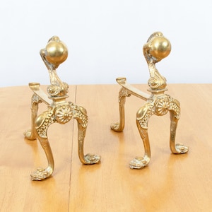 Puede incluir: Un par de soportes de chimenea de metal dorado ornamentado con un remate de bola decorativo en la parte superior. Los soportes tienen un diseño de desplazamiento y se apoyan en tres patas.