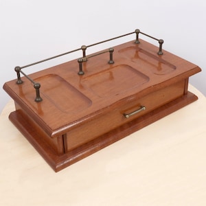May include: A rectangular wooden desk organiser with a polished brown finish. The top surface features three recessed compartments and a thin brass railing. A single drawer is positioned at the front, equipped with a small, horizontal brass handle.