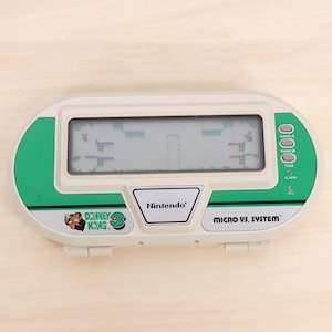 May include: A vintage Nintendo Micro vs. System Donkey Kong 3 handheld game. The device is white and green with a digital screen displaying a game scene. The game has buttons for Game A, Game B, Time, Alarm, and ACL.