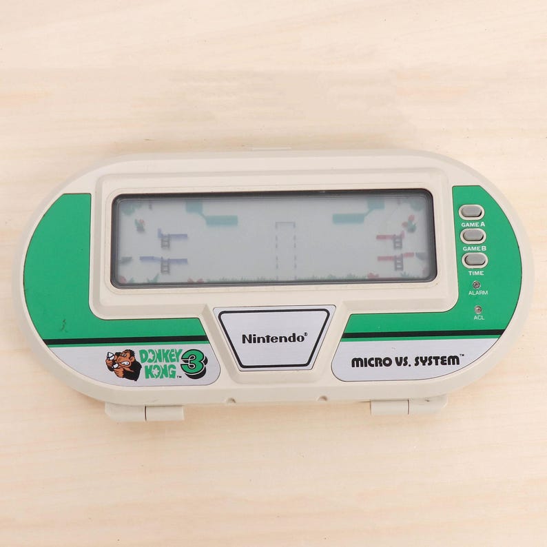 May include: A vintage Nintendo Micro vs. System Donkey Kong 3 handheld game. The device is white and green with a digital screen displaying a game scene. The game has buttons for Game A, Game B, Time, Alarm, and ACL.