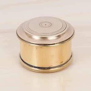 Op de afbeelding: Een kleine, ronde, antieke messing doos met een zilverkleurig deksel. Het deksel heeft een gedetailleerd cirkelvormig ontwerp. De doos heeft een cilindrische vorm met een decoratieve rand rond de basis en het deksel. Het messing heeft een warme, gouden tint.