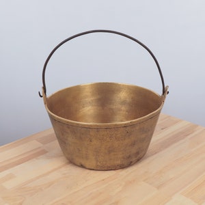 May include: A brass bucket with a dark metal handle. The bucket has a rounded shape and a slightly aged appearance, suggesting it is an antique. The handle arches over the top, providing a way to carry the bucket. The bucket sits on a light wood surface.