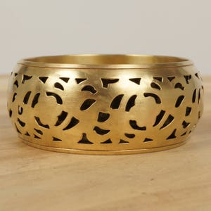 Könnte beinhalten: Ein goldfarbenes Metall-Armreif-Armband mit einem dekorativen Ausschnittmuster. Das Design zeigt ein sich wiederholendes Muster aus geschwungenen Linien und Formen.