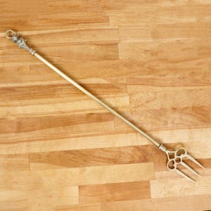 Op de afbeelding: Een lange, messingkleurige haardgereedschap met een decoratieve handgreep en een drietandige vork aan het uiteinde. De handgreep heeft sierlijke details en een ring aan de bovenkant. Het gereedschap staat tegen een houten achtergrond.