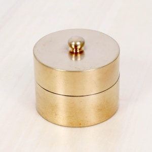 Puede incluir: Un recipiente cilíndrico de latón con un acabado liso y pulido, que presenta un pequeño pomo redondeado centrado en la tapa. El recipiente consta de dos secciones apiladas, con un tono dorado metálico y un diseño circular sencillo.