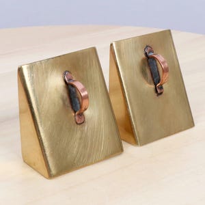 May include: A pair of brass bookends with copper handles. Each bookend is a rectangular prism shape, angled slightly. The brass has a brushed finish, and the copper handles are riveted to the top. They sit on a light-coloured wooden surface.
