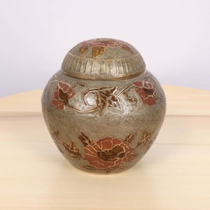 May include: A gray ceramic jar with a lid decorated with gold and red floral designs. The jar has a textured surface and a round shape.