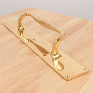 Puede incluir: Una manilla de puerta de latón pulido con un diseño curvo. La manilla está montada en una placa rectangular.