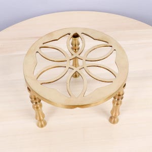May include: A gold-colored metal trivet with a decorative floral design. The trivet has a round top with cut-out patterns and three short, ornate legs. It is designed to protect surfaces from hot items.