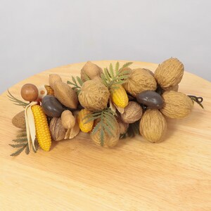 May include: A decorative garland made of nuts, corn, and greenery. The garland features a variety of nuts, including walnuts, almonds, and pecans, as well as small ears of corn and green leaves.
