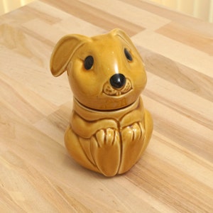 May include: A ceramic honey pot shaped like a brown rabbit with black eyes and a black nose. The rabbit is sitting with its paws crossed.