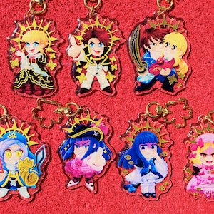 Peut inclure: Ensemble de dix breloques de personnages d'anime colorés avec des accents dorés. Chaque breloque représente un personnage différent avec un halo et un fond en forme d'étoile. Les breloques sont attachées à des chaînes dorées.