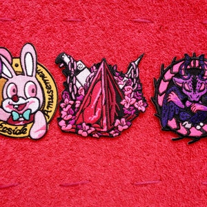 Può includere: Tre toppe ricamate da stirare. La prima toppa è un coniglio rosa che tiene un palloncino viola con il testo "Lakeside Amusements". La seconda toppa è un cristallo rosa e rosso con fiori rosa. La terza toppa è un demone viola con ali nere in un cerchio rosa con spine nere.