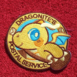 Puede incluir: Parche bordado con un personaje Dragonite amarillo con detalles azules y marrones. El parche dice "DRAGONITE'S POSTAL SERVICE" en un diseño circular. El parche está sobre un fondo rojo texturizado.
