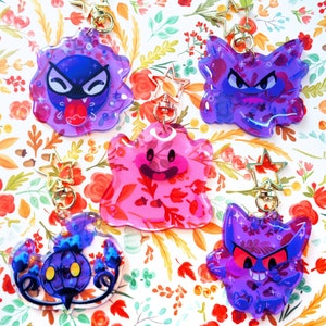 Może przedstawiać: Pięć kolorowych breloków akrylowych z postaciami z kreskówek-duchów. Breloki są fioletowe, różowe i niebieskie, ze złotymi zapięciami w kształcie gwiazd. Tło to kwiatowy wzór w jesiennych kolorach.