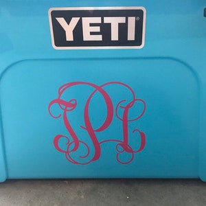 Puede incluir: Una nevera Yeti azul con un monograma rosa de las letras "PL" en la parte delantera.