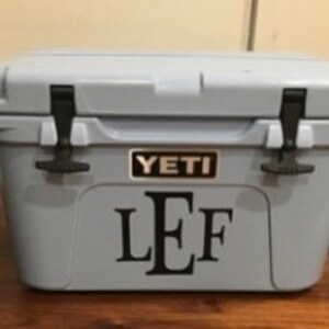 Puede incluir: Una nevera Yeti azul claro con un monograma negro "LEF" en la parte delantera.