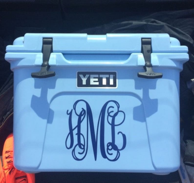 Puede incluir: Una nevera Yeti azul claro con una calcoman&iacute;a monogramada de las letras "HML" en azul oscuro. La nevera tiene asas negras y un logotipo Yeti plateado.