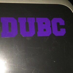 Op de afbeelding: Een paarse sticker met de tekst "DUBC" op een zilveren oppervlak.