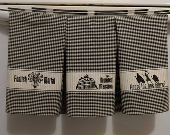 May include: Three black and white checkered kitchen towels with a black and white graphic design. The first towel says "Foolish Mortal" with a silhouette of a ghost. The second towel says "The Haunted Mansion" with a silhouette of the Haunted Mansion. The third towel says "Room for one more?" with a silhouette of three ghosts.