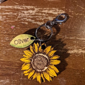 May include: A wooden sunflower keychain with a green leaf tag that says "Oliver" attached to a silver metal ring and clasp.