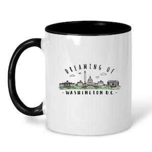 May include: White ceramic coffee mug with a black rim and handle. The mug features a black and white illustration of the Washington D.C. skyline with the text "Dreaming of - Washington D.C. -" printed on the front.