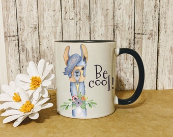 May include: White ceramic mug with a black rim and handle. The mug features a cartoon illustration of a llama with a blue and purple mane and a floral design. The text "Be cool!" is printed in black on the mug.