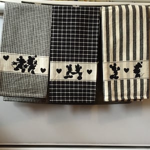 Pode incluir: Três toalhas de cozinha com padrões de xadrez e listras em preto e branco. Cada toalha apresenta uma silhueta preta do Mickey Mouse e da Minnie Mouse de mãos dadas com corações.