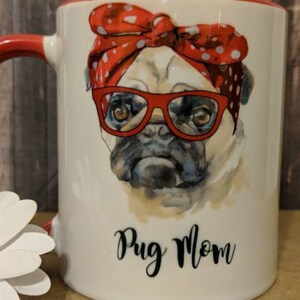 Peut inclure: Mug en c&eacute;ramique blanche avec une anse et un bord rouges. La tasse pr&eacute;sente une illustration &agrave; l'aquarelle d'un carlin portant des lunettes rouges et un bandana &agrave; pois rouges. Les mots "Pug Mom" sont &eacute;crits en script noir.