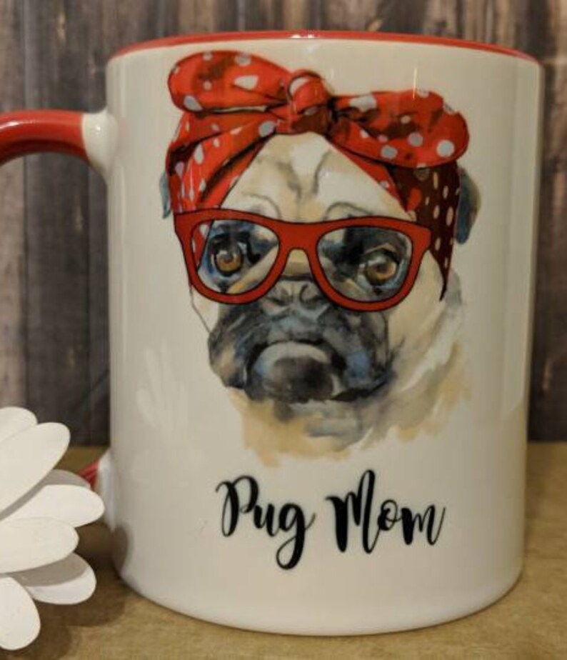 Peut inclure: Mug en c&eacute;ramique blanche avec une anse et un bord rouges. La tasse pr&eacute;sente une illustration &agrave; l'aquarelle d'un carlin portant des lunettes rouges et un bandana &agrave; pois rouges. Les mots "Pug Mom" sont &eacute;crits en script noir.
