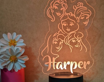Könnte beinhalten: Ein weißes Nachtlicht mit einer durchsichtigen Acrylscheibe, auf der fünf Prinzessinnen und der Name "Harper" abgebildet sind. Das Nachtlicht hat einen schwarzen Sockel mit einem USB-Anschluss.